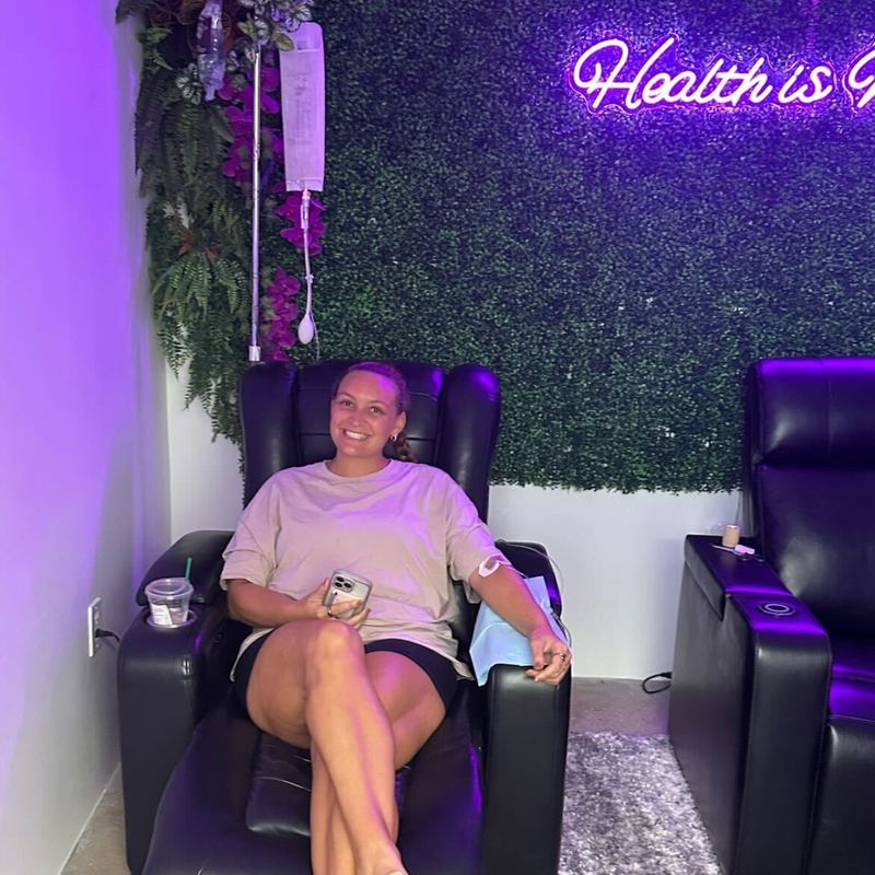 A client smiling in a recliner under the Health is Wealth neon sign