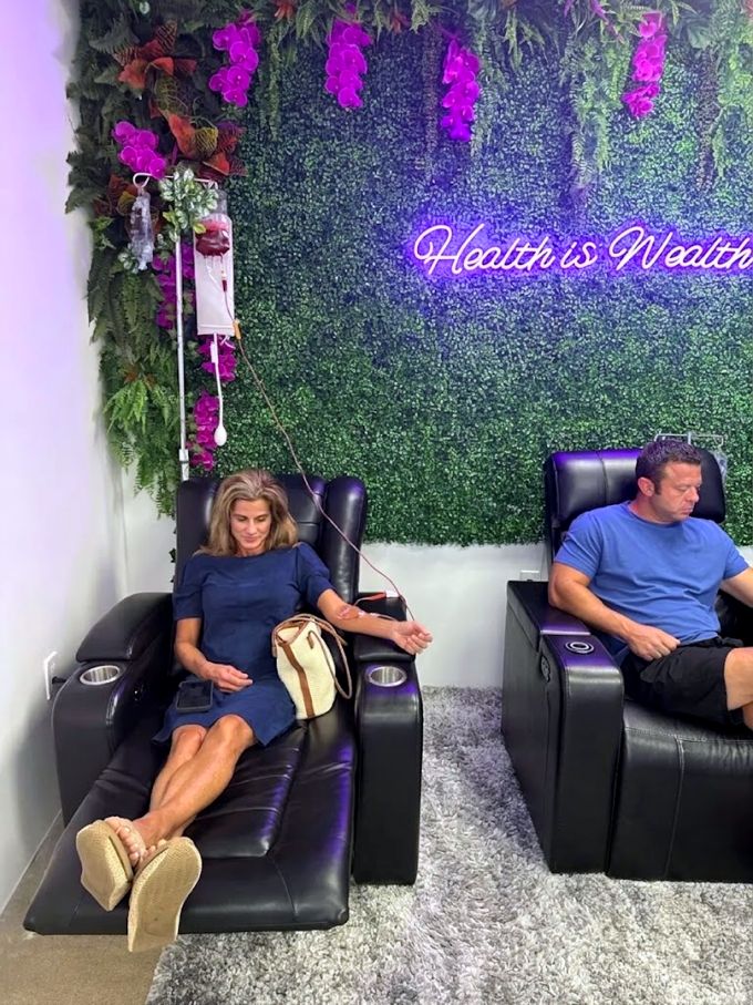 Two clients relaxing in recliners under the Health is Wealth neon sign at the Plano studio