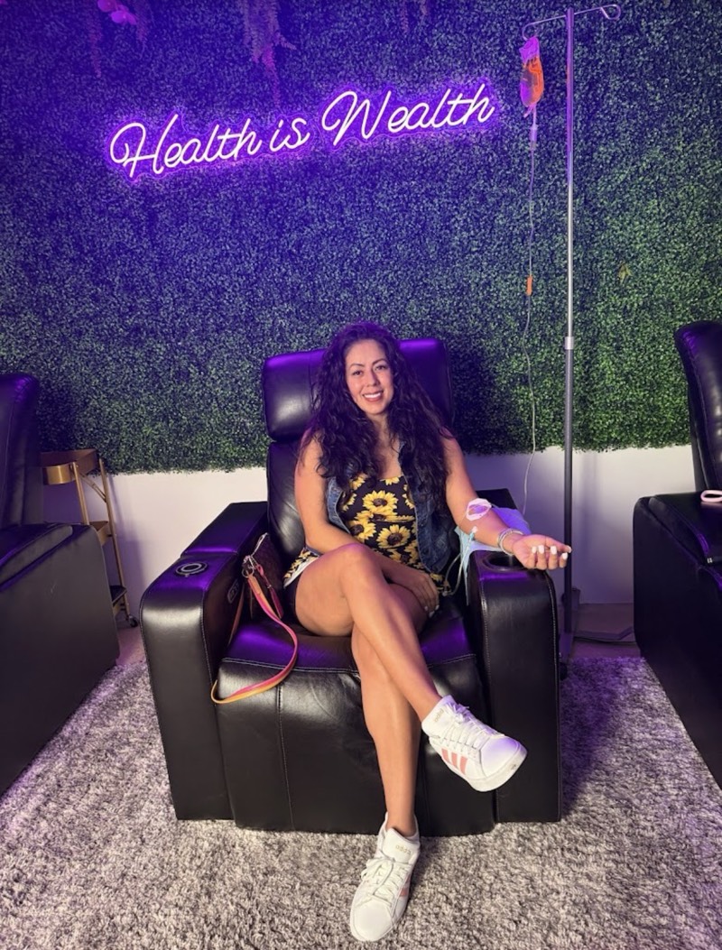 A client smiling in a black recliner under the Health is Wealth neon sign at the Plano studio, getting an IV